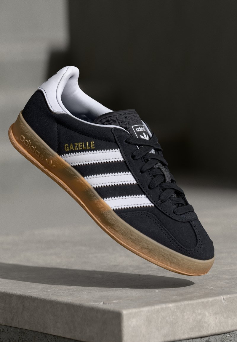 Baskets en daim noir avec des rayures blanches, texte doré "Gazelle" et une semelle en caoutchouc couleur gomme. Design épuré avec un profil bas et un laçage.