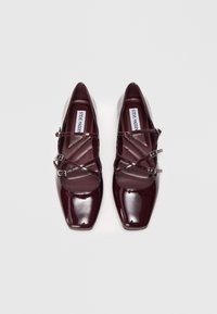 Mirdzošas bordo dzīvokļu kurpes ar kvadrātām purngalām un dubultām sprādzēm, ar uzrakstu Steve Madden, attēlotas no augšpuses uz balta fona.