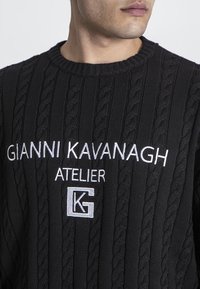 Svart stickad tröja med kabelmönster, rund halsringning och vit broderad text som läser "GIANNI KAVANAGH ATELIER" med en logotyp.