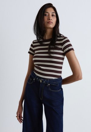 Jeune femme aux longs cheveux foncés portant un t-shirt rayé marron et blanc et un jean taille haute bleu foncé avec une ceinture, debout devant un fond uni.