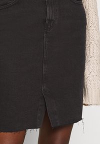 Falda vaquera negra con dobladillo deshilachado y abertura frontal, presenta un corte recto y costura tonal. El material parece suave con una ligera textura.