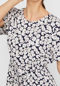 Woman wearing a navy dress with white floral print and short sleeves, tied at the waist with a matching fabric belt.