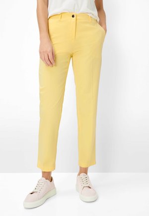 Dame bukser i gul, slim fit med frontknap og lynlås, parret med lyserøde sneakers, stående mod en hvid baggrund.