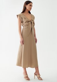 Robe en satin beige avec un décolleté en V profond, des manches courtes et une taille nouée. Présente une silhouette fluide et une texture lisse. Associée à des sandales beiges.