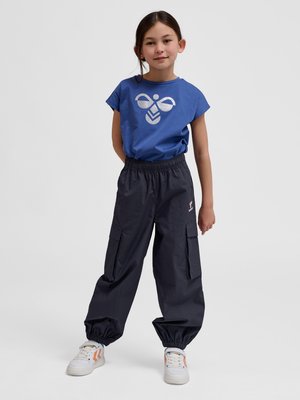 Młoda dziewczyna stoi z rękami za plecami, ubrana w niebieską koszulkę z białym logo pszczoły, czarne spodnie cargo i białe tenisówki z pomarańczowymi detalami.