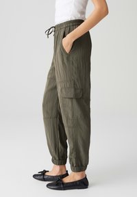 Olijfgroene, lichte cargobroek met een elastische tailleband en boorden, uitgerust met zijzakken en een gestructureerde stof. Gecombineerd met zwarte platte schoenen.