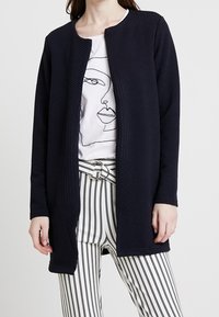 Marinblå ribbad kappa med rund halsringning, öppen front och strukturerade paneler, kombinerad med en vit t-shirt med grafiskt tryck och randiga byxor.