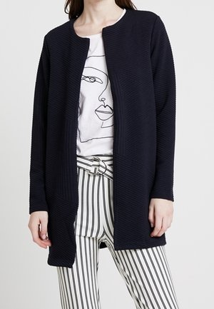 Marineblauwe geribde cardigan met ronde halslijn, open voorkant en textuurdetails, gecombineerd met een witte graphic tee en gestreepte broek.