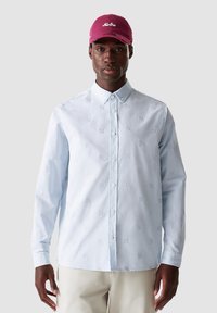 Camicia button-up azzurro chiaro con motivi ricamati, maniche lunghe e colletto a punta, indossata con un cappellino da baseball bordeaux.