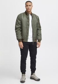 Olive green bomber jacket with a zipper front, ribbed cuffs, and stand collar. Paired with black pants and gray athletic shoes.