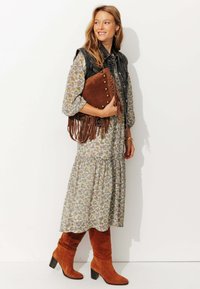 Robe florale avec un ourlet à volants, gilet en denim noir, sac à franges marron avec des clous dorés, et bottes en daim orange hauteur genou avec des talons carrés.