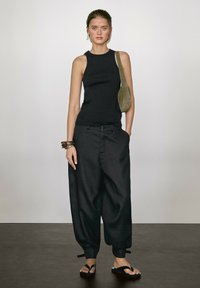 Black sleeveless top paired with loose, dark gray pants. The model carries a green handbag and wears black sandals with straps.