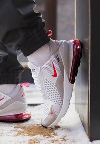 Vit idrottssko med röda accenter, ovandel i mesh, vadderad sula och en genomskinlig röd luftenhet; detaljerad textur och mönster.