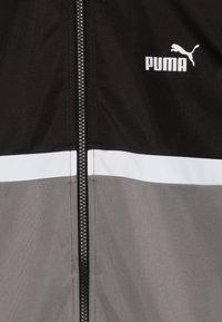 Sort og grå zip-up jakke med en hvid vandret stribe og Puma-logo. Glat stof med et slankt design og kontrasterende farver.