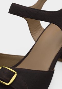 Κοντινό πλάνο καφέ σουέτ ψηλοτάκουνα σανδάλια με χρυσή αγκράφα και μπεζ επενδεδυμένη εσωτερική σόλα.