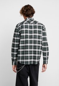 Vihreä-valkoruudullinen flanellipaita, jossa pitkät hihat, napilliset hihansuut ja takana oleva yoke-yksityiskohta. Päällä mustat housut ja ketjusomisteet.