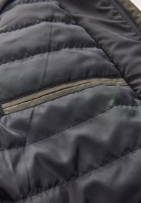 Gros plan sur un tissu noir matelassé avec des coutures horizontales et une ouverture de poche étroite de couleur vert olive.