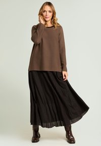 Pull marron à coupe décontractée, jupe longue noire plissée, et bottes montantes marron foncé. Texture lisse, design en couches, et éclat subtil.