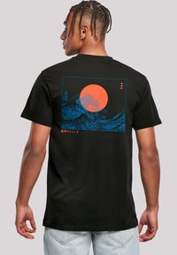 Black cotton t-shirt featuring a blue graphic design of waves and a bright orange sun on the back, with Japanese text below the illustration.