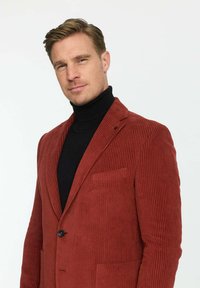 Rote Cordjacke mit vertikalen Rippungen, Vorderseite mit einem Knopf und einer Brusttasche, getragen über einem schwarzen Rollkragenpullover.