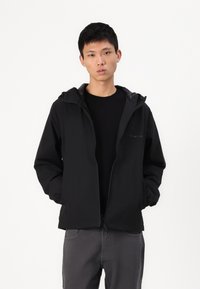 Svart hettegenser laget av lett materiale, med glidelås foran, to sidelommer og en teksturert innerside. Modellen har på seg grå bukser.