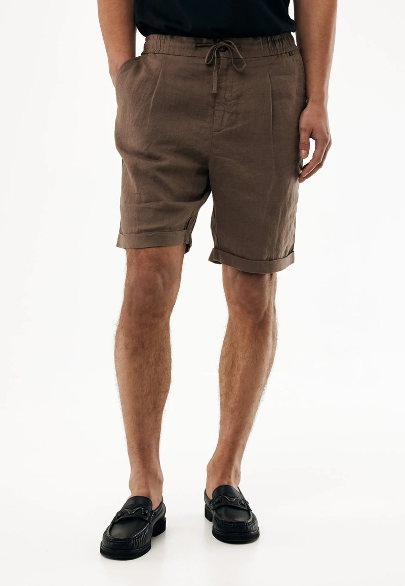 Mand iført brune shorts med snøre og opbrettede kanter samt sorte lædersko, stående mod en ensfarvet hvid baggrund.