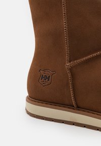 Bota de ante marrón con costuras visibles y un parche de logo. Presenta una suela de color crema con textura antideslizante y un diseño de punta redonda.