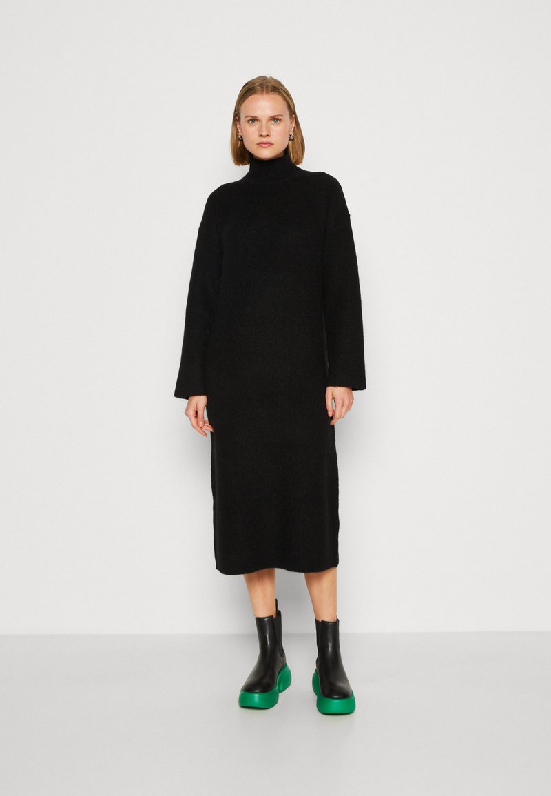 Vestito nero a maniche lunghe con collo alto, realizzato in un materiale morbido, caratterizzato da una vestibilità comoda. Abbinato a stivaletti neri chunky con suola verde.