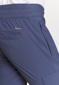 Námořnické modré turistické kalhoty vyrobené z měkké látky, s elastickým pasem, zipovou kapsou a malou štítkovou nášivkou.