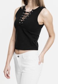 Černý bezrukávový crop top s šněrováním na přední straně, obsahující kovové očička. V kombinaci s bílými džíny, které ukazují obepínající siluetu.