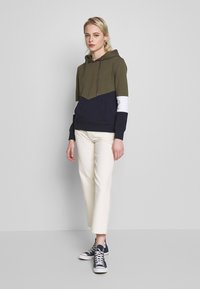 Sweatshirt com capuz em blocos de cores verde-oliva, azul-marinho e branco; apresenta um bolso frontal e capuz com cordão. Combinado com calças creme e tênis.