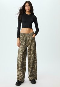 Donna con lunghi capelli scuri, che indossa un crop top nero a maniche lunghe, pantaloni larghi con stampa leopardata e scarpe con piattaforma nere, in piedi con una mano in tasca.