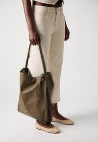 Pessoa a usar jeans cropped creme e sapatilhas bejes, segurando uma grande mala de pele verde-azeitona com detalhes em dourado.