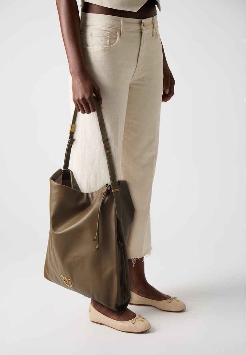 Pessoa a usar jeans cropped creme e sapatilhas bejes, segurando uma grande mala de pele verde-azeitona com detalhes em dourado.