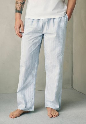 Férfi világoskék és fehér függőlegesen csíkozott pizsanadrágban, kezét a zsebében tartva, mezítláb áll szürke padlón, szürke fal előtt.