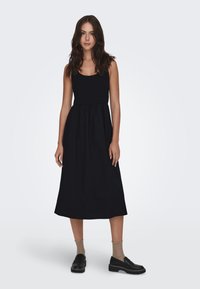 Mustat midi-mekko, jossa on istuva yläosa, kerätty vyötärö ja valuva hame. Mallilla on päällä mustat loafers-kengät ja teksturoidut, vaaleansävyiset sukat.