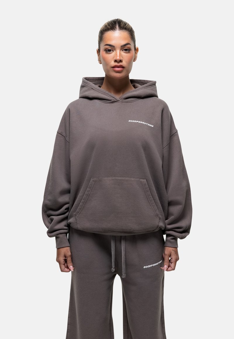 Felpa oversize marrone scuro con tasca frontale, maniche a costina e testo "GOODFORNOTHING". Abbinata a pantaloni larghi coordinati.
