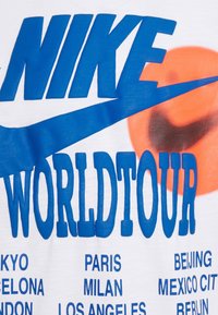 Valkoinen puuvillapaita, jossa on rohkea sininen "NIKE" ja "WORLDTOUR" -teksti, ja kaupunkien nimet painettuna alle. Suuri oranssi grafiikka täydentää muotoilua.