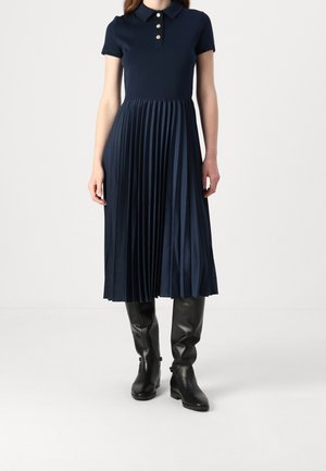 Frau trägt ein marineblaues Polokleid mit kurzen Ärmeln, einem plissierten Midirock und schwarzen kniehohen Lederstiefeln.