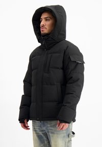 Zwarte pufferjack met capuchon, voorzien van een ritssluiting, twee grote voorzakken en een zak op de linkermouw. Glad, gewatteerd textuur.