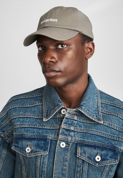 Khaki baseball cap with white embroidered text, worn with a blue denim jacket featuring vertical stitching and large buttoned chest pockets.