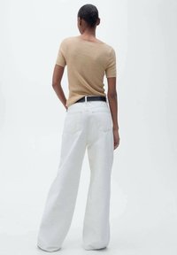 Persona de pie de espaldas, vistiendo una camiseta de manga corta de canalé beige metida en unos pantalones blancos de pierna ancha de cintura alta con cinturón negro.