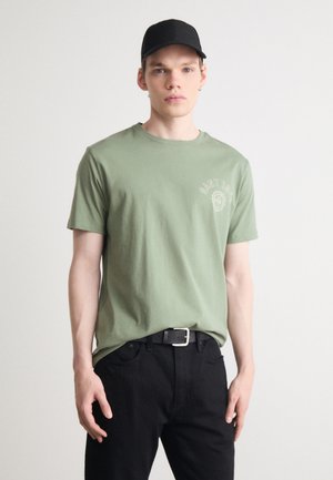 Olive green cotton t-shirt with a crew neck and short sleeves, featuring a subtle logo print on the left chest. Worn with black jeans.