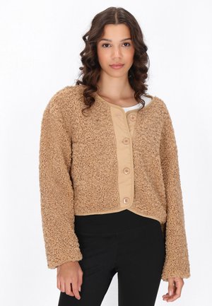 Jonge vrouw met krullend haar, die een beige gestructureerde jas met knopen draagt en een zwarte broek, staand tegen een effen witte achtergrond.
