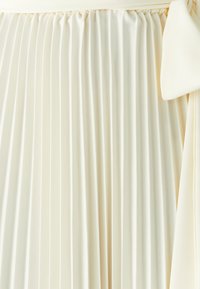 Plissierter Rock aus cremefarbener Stoff mit einer weichen Textur; verfügt über einen elastischen Bund und ein gebundenes Akzentdetail auf einer Seite.