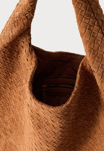 Καφέ τσάντα ώμου από σουέντ με πλεκτή υφή, φαρδιές λαβές και εμφανές εσωτερικό φερμουάρ, πάνω σε λευκό φόντο.