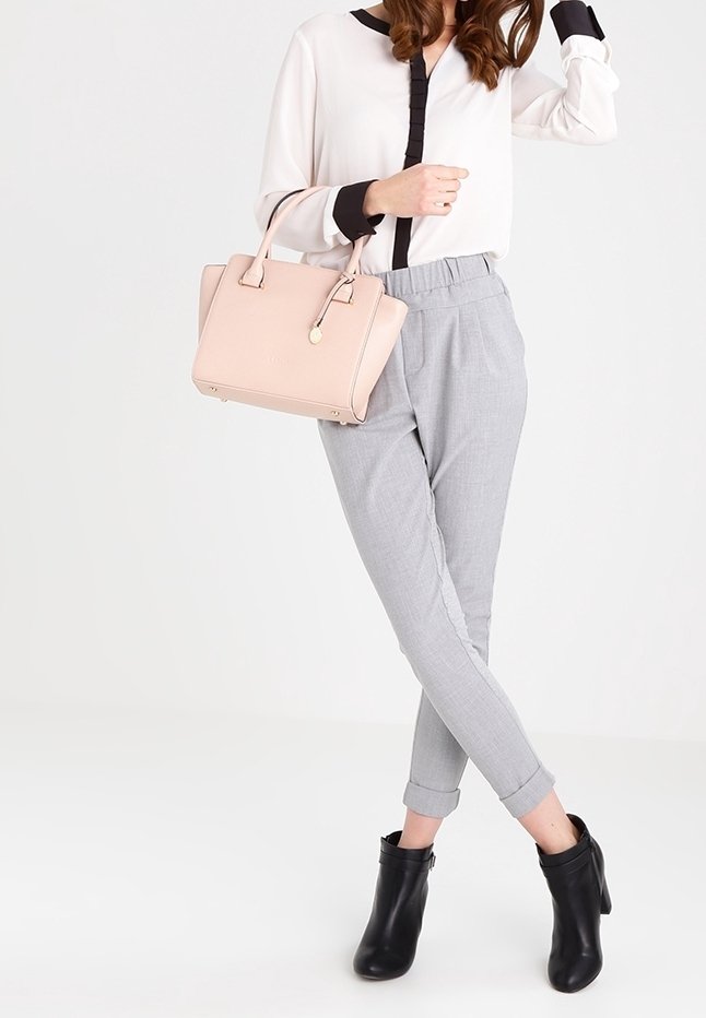 Femme portant un chemisier blanc avec un liseré noir, un pantalon gris retroussé, des bottines noires, et tenant un sac à main rose clair sur son bras.