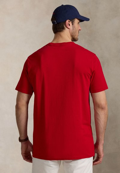 Red cotton t-shirt with short sleeves and a crew neck, featuring a smooth texture. A navy cap is worn, positioned at the back.