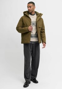 Olivgrüne Kapuzenjacke über einem cremefarbenen Pullover mit gemusterten Akzenten, graue Hose und schwarze Schuhe. Die Jacke hat einen Reißverschluss.