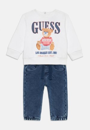 Sweat-shirt blanc avec imprimé d'ours et logo "GUESS", associé à un pantalon en denim bleu foncé avec taille élastique et ourlets retroussés.